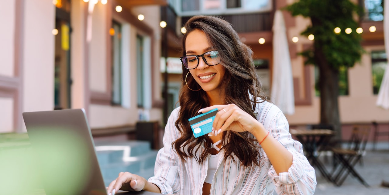 A woman in glasses sits at a table and smiles as she holds her payment card and uses her laptop.